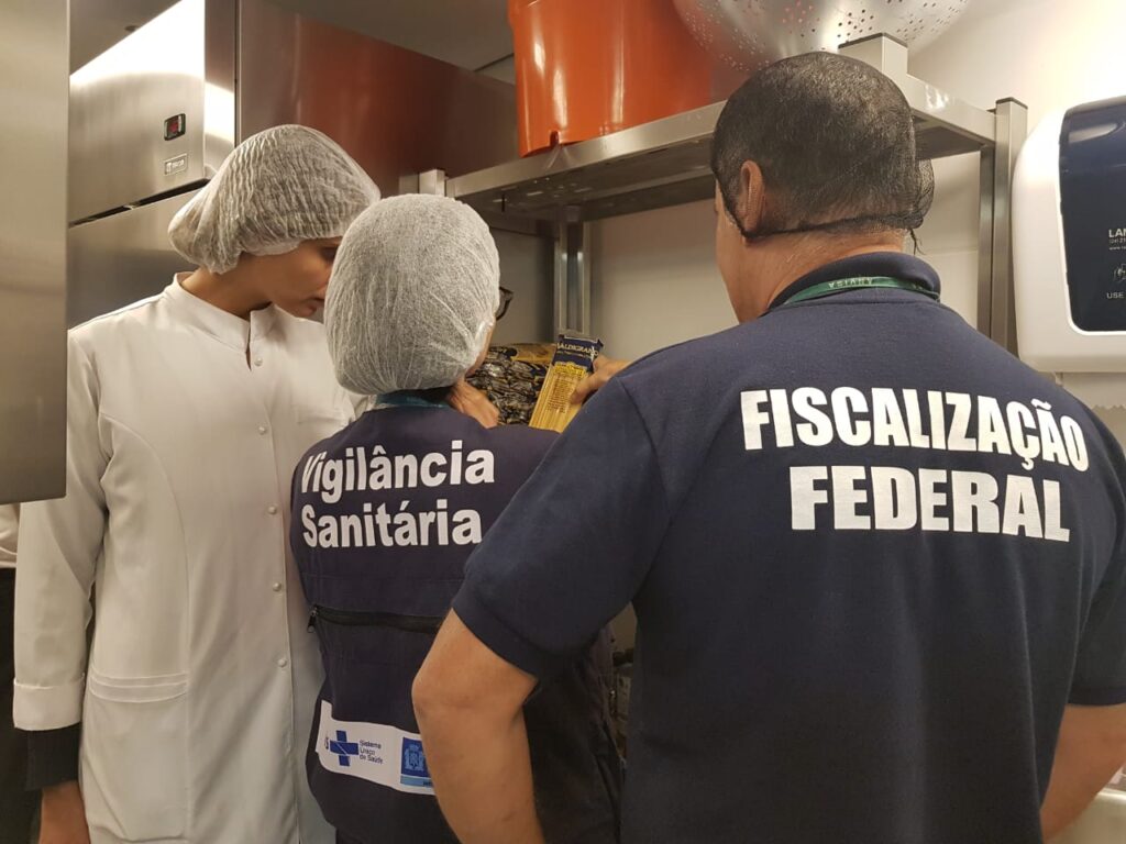 Fiscais da Vigilância Sanitária e da Fiscalização Federal inspecionam a cozinha de um restaurante, acompanhados por um funcionário de jaleco branco, reforçando a importância da higiene e da desratização em ambientes alimentícios.